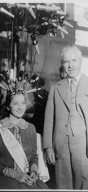 Woman with hair in curling machine sits next to man standing